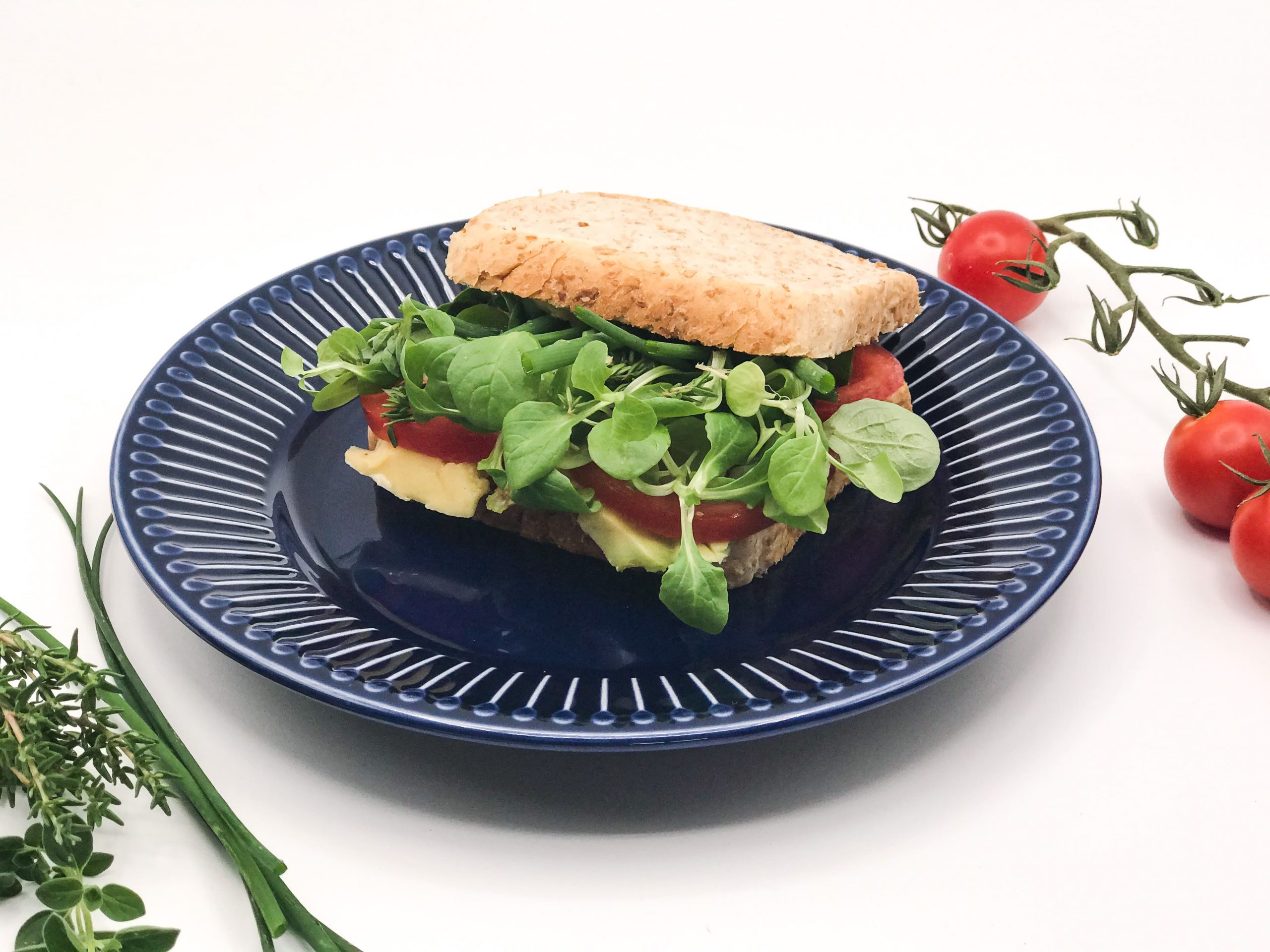 Ein Teller, darauf liegt ein Sandwich, neben Teller ein paar Tomaten am Stiel und ein paar Kräuter.