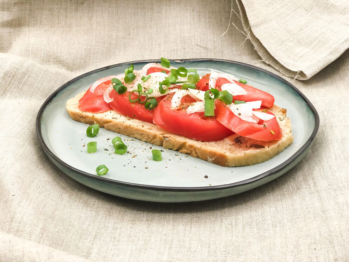 Auf einem Teller: Eine Scheibe Bauernbrot, darauf die Tomatenscheiben, Zwiebeln und Frühlingszwiebeln.